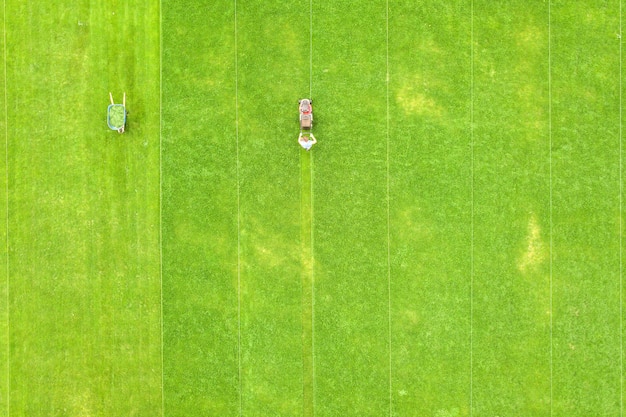 Vista aérea de la pequeña figura de trabajador hombre recortando pasto verde con mashine de corte en el campo del estadio de fútbol en verano.