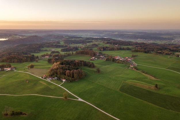 Vista aérea de pequeñas casas de campo dispersas con techos de tejas rojas entre campos de cultivo verdes y bosque de montaña distante en verano.