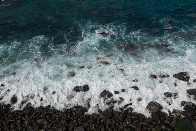 Vista aérea de la playa negra con olas salpicando rocas volcánicas Océano azul turquesa brillante Fotografía de la costa Vista superior de la orilla del océano Patrón blanco de espuma de mar sobre piedras Foto marina