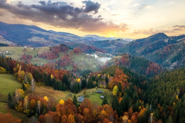 Vista aérea del pueblo distante con pequeñas casas de pastor en prados de colinas anchas entre árboles de bosque otoñal en las montañas de los Cárpatos ucranianos al atardecer.