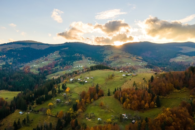 Vista aérea del pueblo distante con pequeñas casas de pastor en prados de colinas anchas entre árboles de bosque otoñal en las montañas de los Cárpatos ucranianos al atardecer.