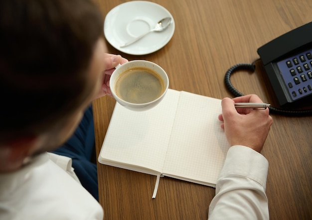 Vista aérea recortada de un empresario que sostiene una taza de cerámica blanca con café recién hecho y toma notas en un cuaderno, sentado en un escritorio con teléfono fijo en la habitación del hotel durante un viaje de negocios