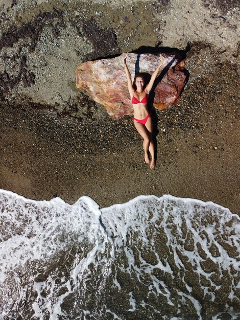 Vista aérea superior em uma jovem loira sensual de biquíni vermelho deitada na rocha perto da água no mar