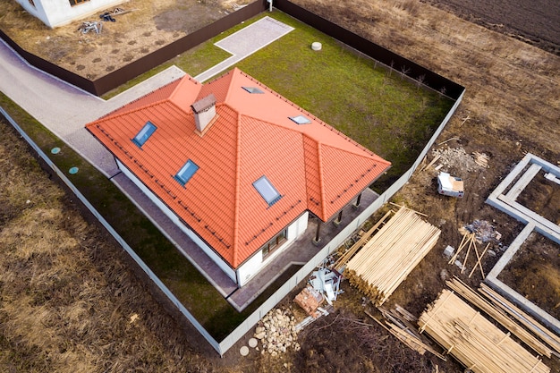 Vista aérea del techo de la casa nueva con ventanas del ático y sitio de construcción, cimientos de la futura casa, pilas de ladrillos y troncos de madera para construcción.