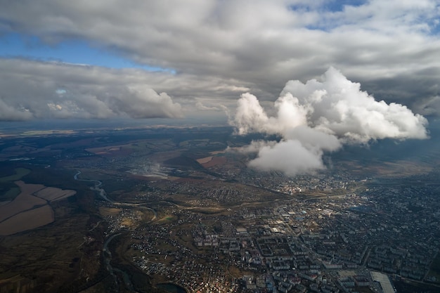 Vista aérea desde la ventana del avión a gran altura de una ciudad distante cubierta de cúmulos hinchados que se forman antes de la tormenta