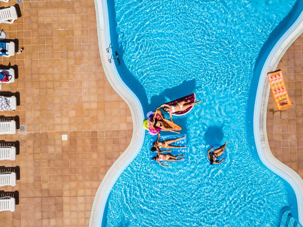 Vista aérea vertical de um grupo de amigas se divertindo nas férias de verão no resort com piscina com infláveis coloridos lilos na água azul