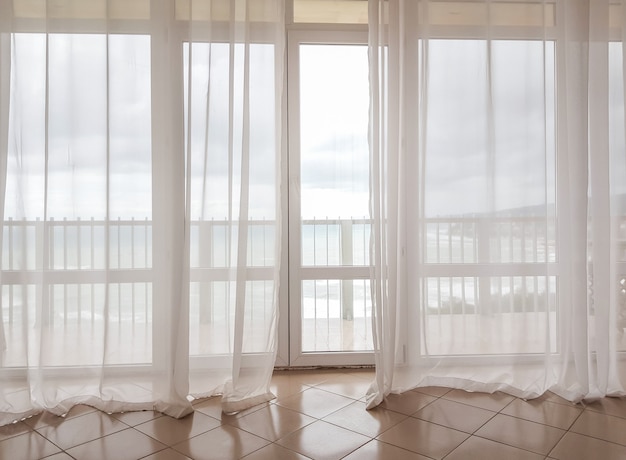 Vista al mar a través de una cortina transparente en una enorme ventana panorámica con balcón, interior moderno de apartamentos en un hotel de lujo.