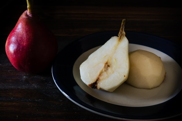 Vista alta de una pera de batalla roja sin pelar a la mitad en un plato y otras peras en la mesa Fondo oscuro Productos orgánicos y naturales alimentos saludables y saludables