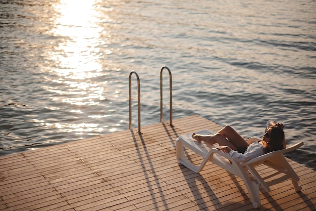 Vista de alto ángulo de una mujer delgada y bronceada relajándose en una silla de terraza en un muelle de madera durante la puesta de sol Una bonita morena disfrutando de vacaciones de verano en la costa del mar Adriático Concepto de recreación