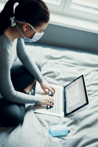 Vista de alto ángulo de una mujer con máscara usando una computadora portátil en la cama