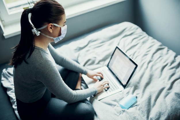 Vista de alto ángulo de una mujer con máscara usando una computadora portátil en la cama