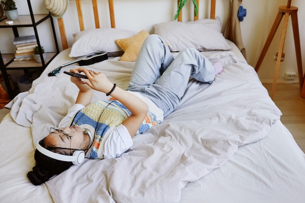 Vista de ángulo alto de una adolescente de moda acostada en la cama y usando un teléfono inteligente en el interior de la habitación acogedora, espacio de copia