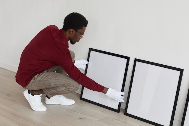 Vista de ángulo alto al hombre afroamericano configurando marcos negros en blanco en el piso mientras planifica una galería de arte o una exposición,