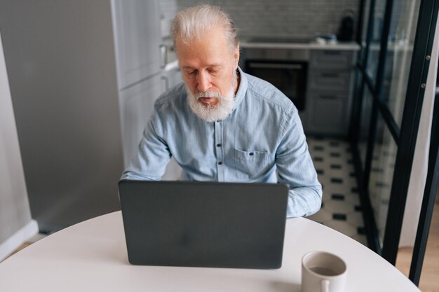 Vista de ángulo alto de un anciano maduro enfocado mirando la pantalla de una computadora portátil trabajando en una computadora sentado en la mesa de la cocina con una taza de café Viejo hombre de negocios escribiendo mensajes resolviendo problemas de negocios