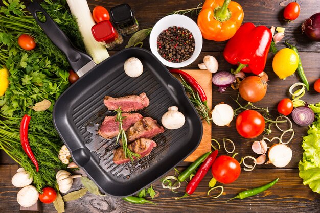 Vista de ángulo alto de carne asada rara en rodajas chisporroteando en una sartén de hierro fundido caliente rodeada de ingredientes frescos