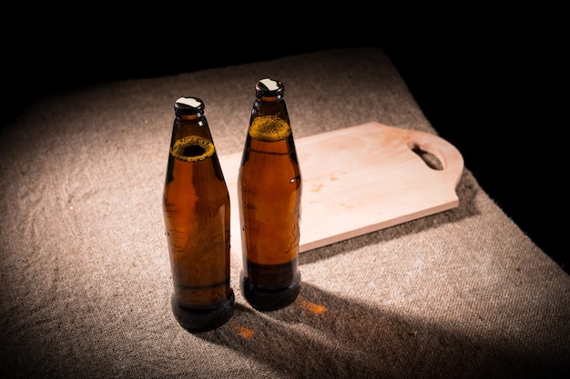 Vista de ángulo alto de dos botellas llenas de cerveza una al lado de la otra en una mesa cubierta de arpillera con una tabla de cortar de madera iluminada desde arriba en un entorno oscuro