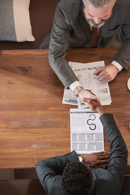 Vista de ángulo alto de dos hombres de negocios sentados en la mesa discutiendo el contrato comercial y dándose la mano para concluir un trato