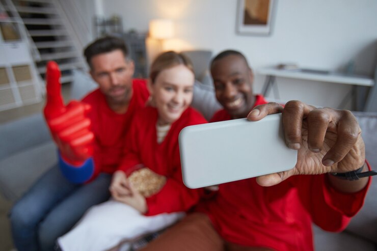 Foto vista de ángulo alto en un grupo de fanáticos de los deportes vestidos de rojo y tomando selfie mientras ve el partido en casa, concéntrese en primer plano