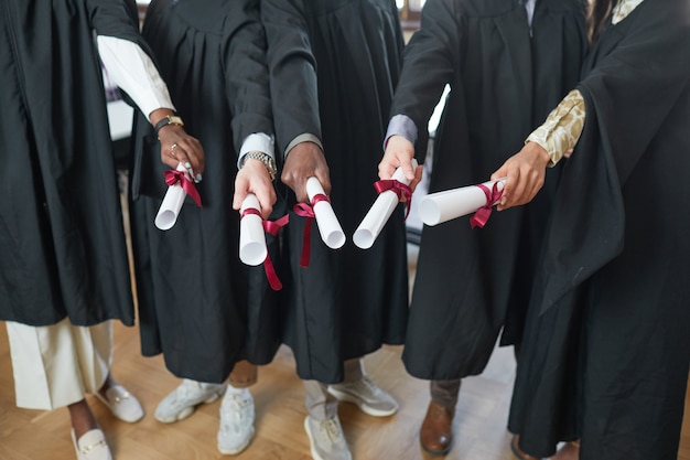Vista de ángulo alto en el grupo multiétnico de jóvenes vistiendo túnicas de graduación y señalando diplomas a la cámara