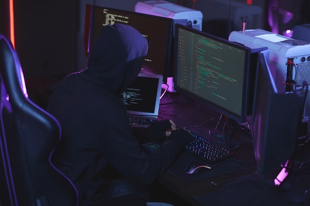 Vista de ángulo alto en hacker de seguridad cibernética irreconocible con capucha mientras trabaja en código de programación en una habitación oscura, espacio de copia