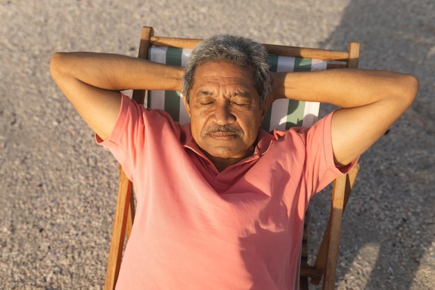 Vista de ángulo alto de un hombre birracial mayor durmiendo con las manos detrás de la cabeza en una silla relajándose en la playa