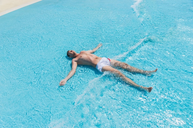 Vista de ángulo alto de hombre joven con traje de baño acostado boca arriba con los brazos y las piernas extendidas en las olas poco profundas de la piscina en un día soleado de verano