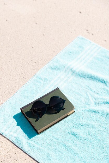 Foto vista de ángulo alto del libro con gafas de sol y toalla azul en la playa de arena durante el día soleado, espacio para copiar. concepto inalterado, verano, protección, hobby, ausencia y vacaciones.