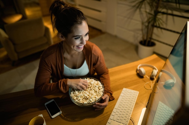 Vista de ángulo alto de mujer feliz relajándose por la noche y comiendo palomitas de maíz mientras ve una película en su computadora en casa