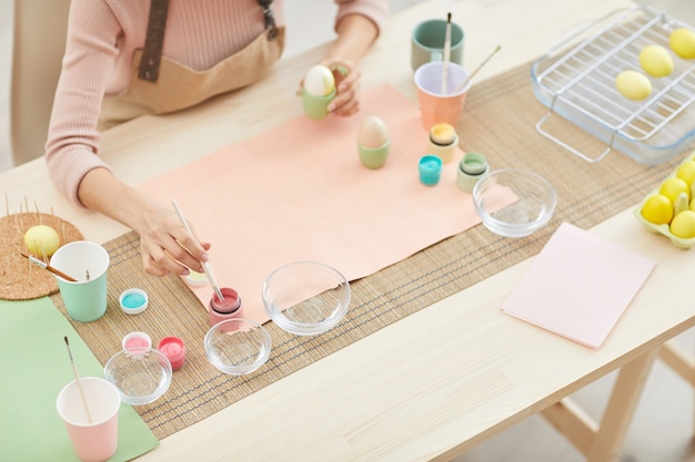 Vista de ángulo alto de mujer irreconocible pintando huevos en colores pastel para la Pascua mientras está sentado a la mesa en la cocina, espacio de copia