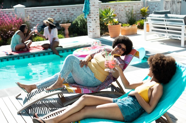 Vista de ángulo alto de mujeres birraciales brindando bebidas mientras disfrutan relajándose junto a la piscina en verano