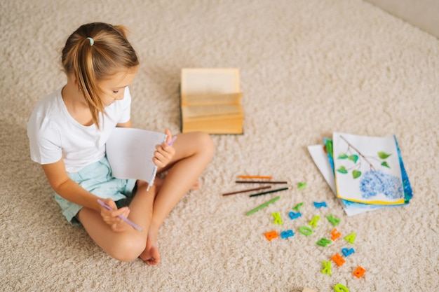 Vista en ángulo alto de una niña bonita dibujando haciendo una foto sentada en el suelo en la habitación de los niños con un interior moderno y luminoso