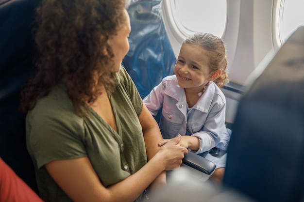 Vista de ángulo alto de niña sentada en el avión, sonriendo a su madre mientras viajan juntos. Familia, concepto de vacaciones