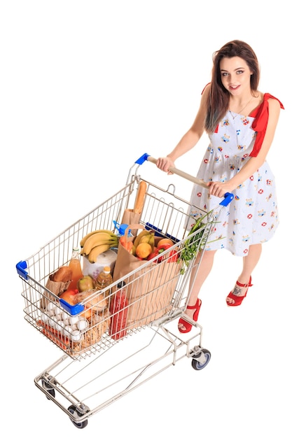 Vista de ángulo alto de niña sonriendo a la cámara mientras empuja un carrito de compras lleno de comestibles aislado sobre fondo blanco.