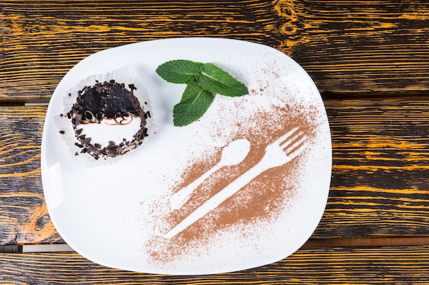 Vista de ángulo alto de postre decadente recubierto de virutas de chocolate y servido en un plato con guarnición de hojas de menta y contornos de utensilios espolvoreados en cacao y descansando sobre la superficie de la mesa de madera