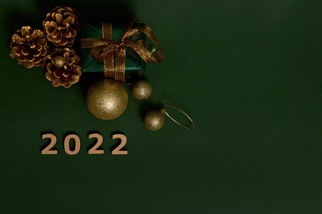 Vista de ángulo alto de un regalo en papel de regalo verde con lazo dorado, conos de pinos, números de madera que simbolizan el año 2022 y juguetes de árbol de Navidad dorados sobre fondo oscuro. Copie el espacio para el anuncio