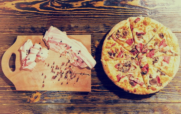 Vista de ángulo alto de sabrosa pizza con variedad de coberturas y queso en la mesa de madera, cerca de la tabla de cortar se encuentra con tocino y especias