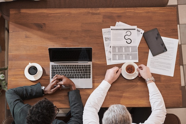 Vista de ángulo alto de socios comerciales sentados en la mesa de madera y tomando café durante el trabajo con documentos y computadora portátil