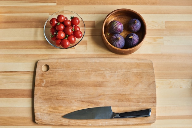 Vista de ángulo alto de tabla de cortar de madera con cuchillo con tomates e higos en los tazones sobre la mesa