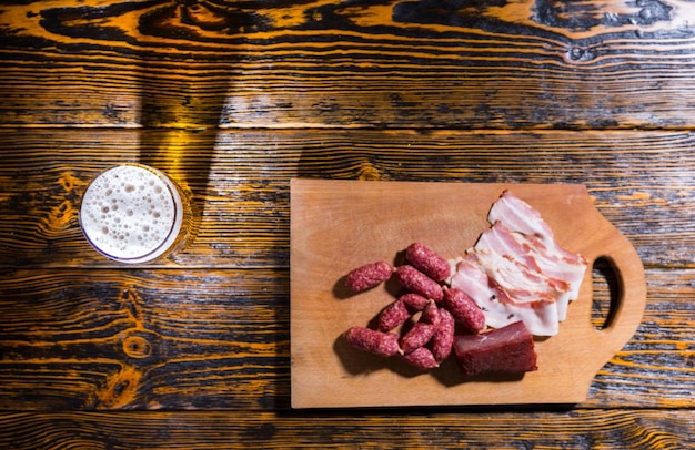 Vista de ángulo alto de la tabla de cortar con salchichas y carne en la mesa de madera cerca de un vaso de cerveza