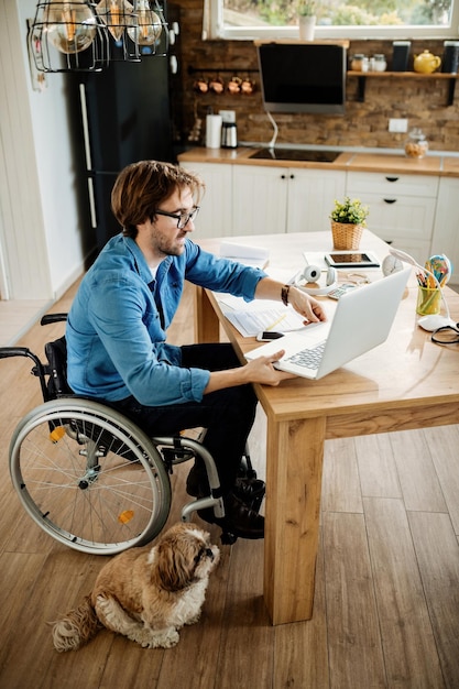 Vista de ángulo alto del trabajador independiente en silla de ruedas usando una computadora portátil y trabajando en casa mientras su perro está con él.