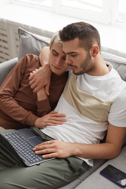 Vista de ángulo alto vertical en dos hombres jóvenes que se abrazan mientras usan la computadora portátil juntos en casa, una pareja gay en la vida cotidiana