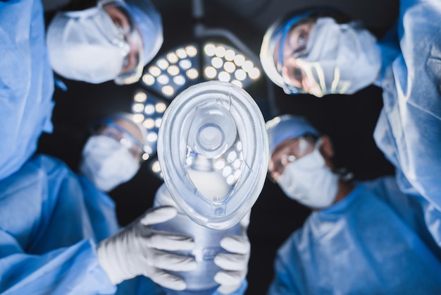 Foto vista de ángulo bajo del anestesista sosteniendo una máscara de oxígeno por encima del paciente en la sala de cirugía con un equipo de profesionales cirujanos
