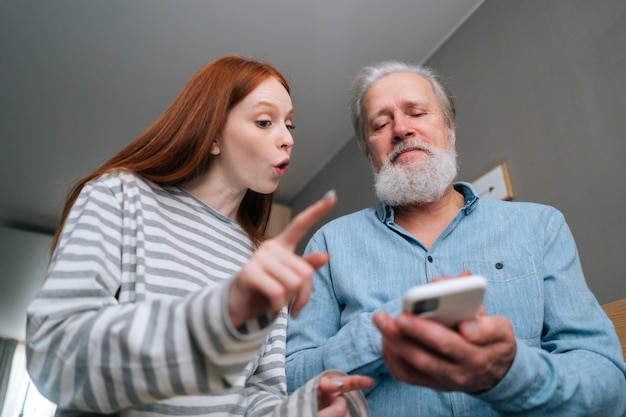 Vista de ángulo bajo del apuesto abuelo mayor barbudo que estudia para usar el teléfono móvil bajo la guía de una joven nieta positiva