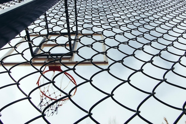 Vista de ángulo bajo del aro de baloncesto visto a través de la valla de cadena contra un cielo despejado