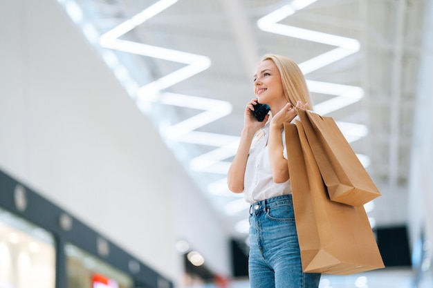 Vista de ángulo bajo de una atractiva joven rubia sonriente con ropa elegante hablando por teléfono móvil sosteniendo bolsas de papel con la compra