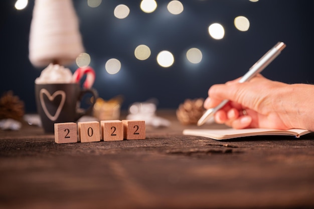 Vista de ángulo bajo del cartel de 2022 en cubos de madera con adornos navideños en el fondo y mano femenina escribiendo planes de año nuevo en un cuaderno.