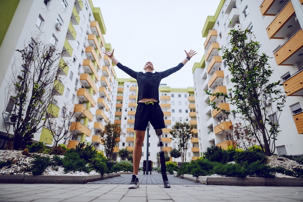 Foto vista de ángulo bajo de deportista guapo con pierna artificial de pie con las manos en el aire al aire libre rodeado de edificios.