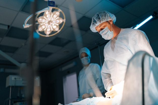 Vista de bajo ángulo del equipo cooperativo de cirujanos que procesan la operación quirúrgica en una sala de operaciones oscura