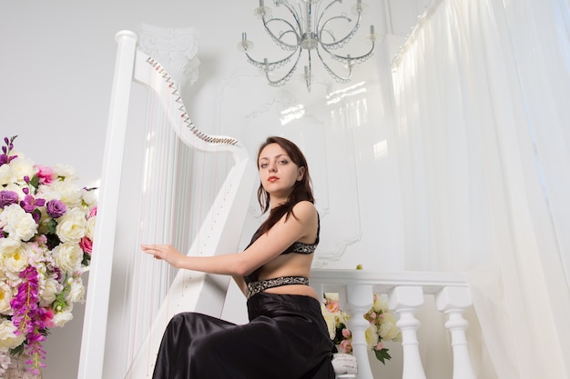 Vista de ángulo bajo de una glamorosa joven tocando el arpa en un recital musical formal en un elegante escenario blanco decorado con un cuenco de flores frescas y cortinas blancas brillantes