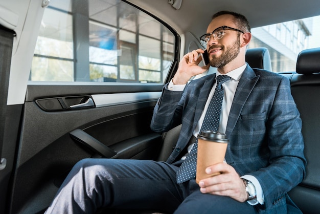 vista de ángulo bajo de un hombre de negocios feliz hablando en teléfono inteligente y sosteniendo una taza de papel en el coche